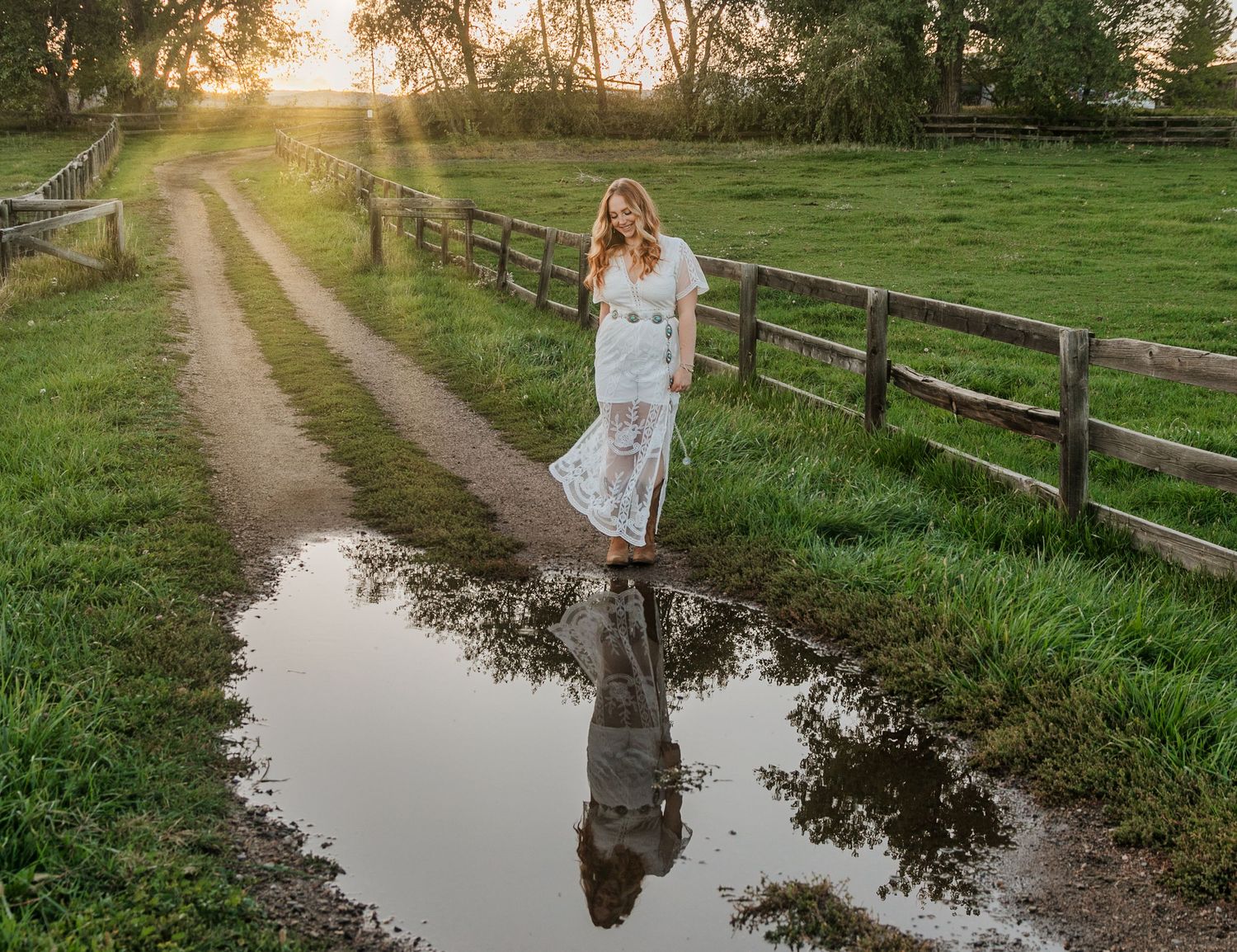 Woman in white dress walks along rustic country path with puddles reflecting trees during golden sunset hour.