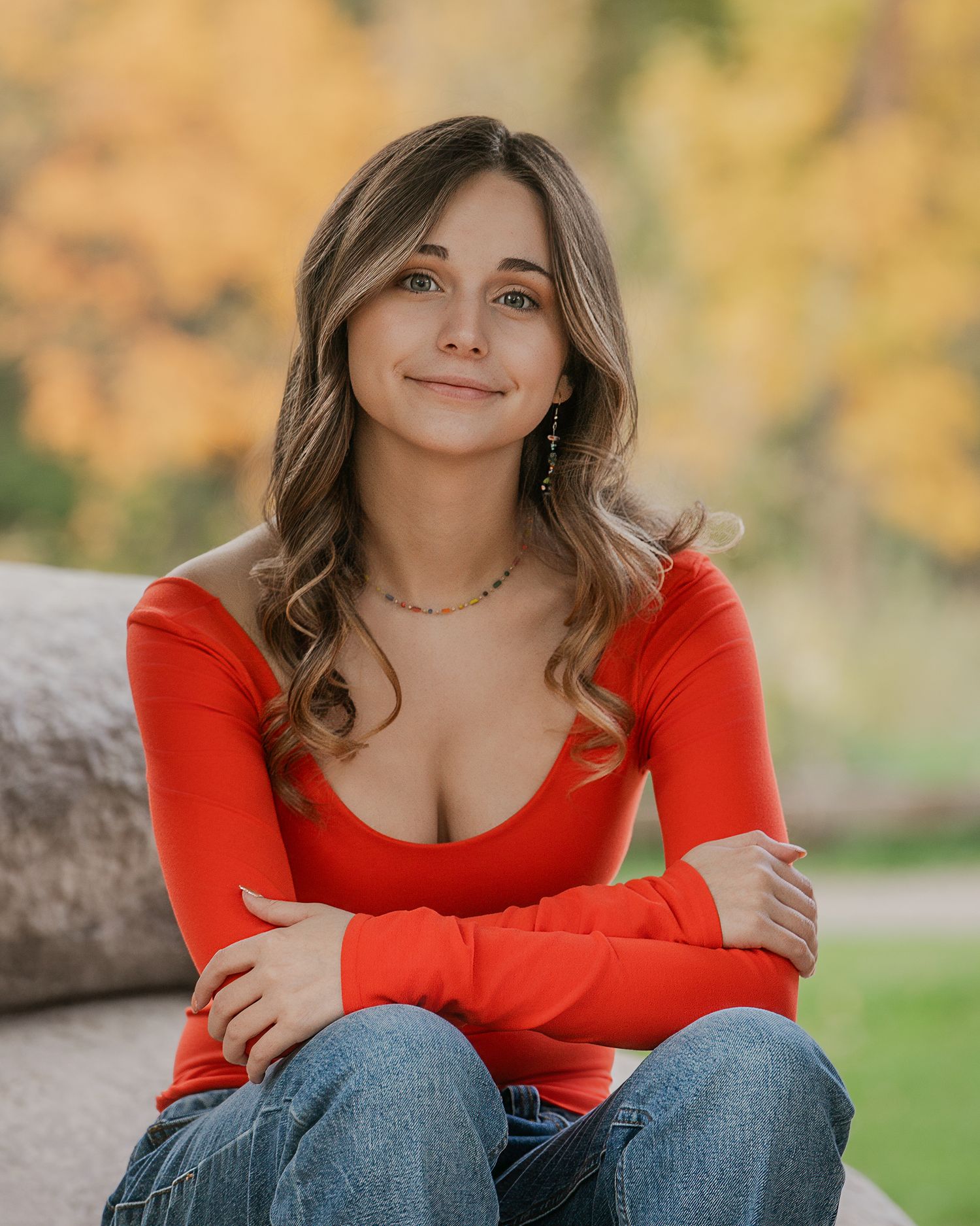 A woman in a red sweater sits outdoors during autumn with golden foliage in the background.