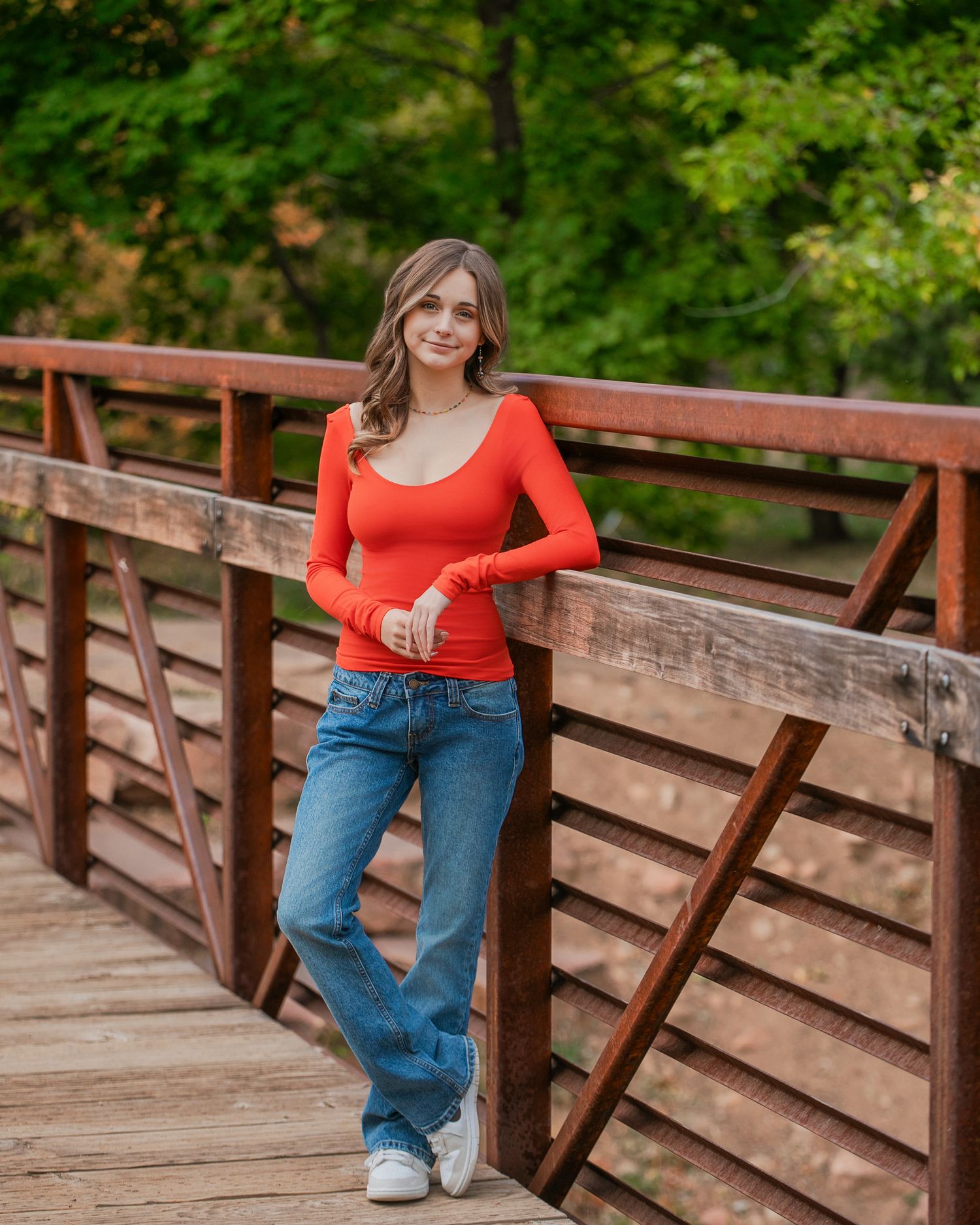A person in a red long-sleeve top and blue jeans poses on a rustic wooden bridge in a lush forest setting.