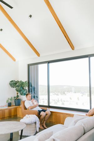 Man sitting in a modern living room chair holding a baby near a large window with scenic views.