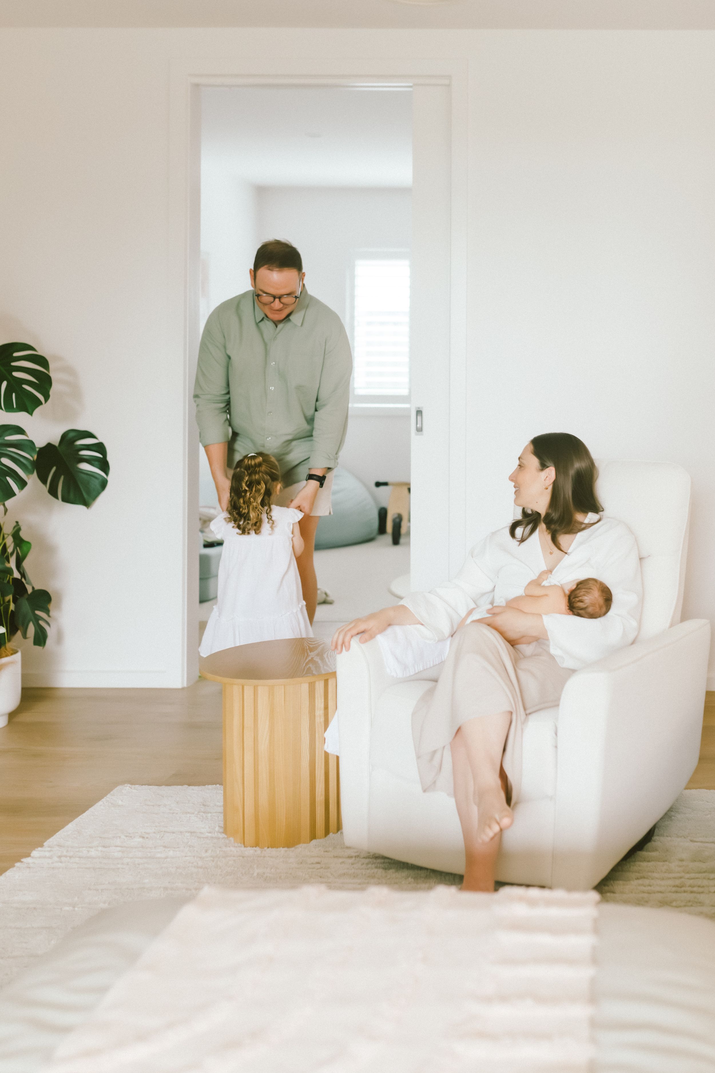 A mother breastfeeds a newborn in a white armchair while her toddler and husband interact nearby in a bright modern home.