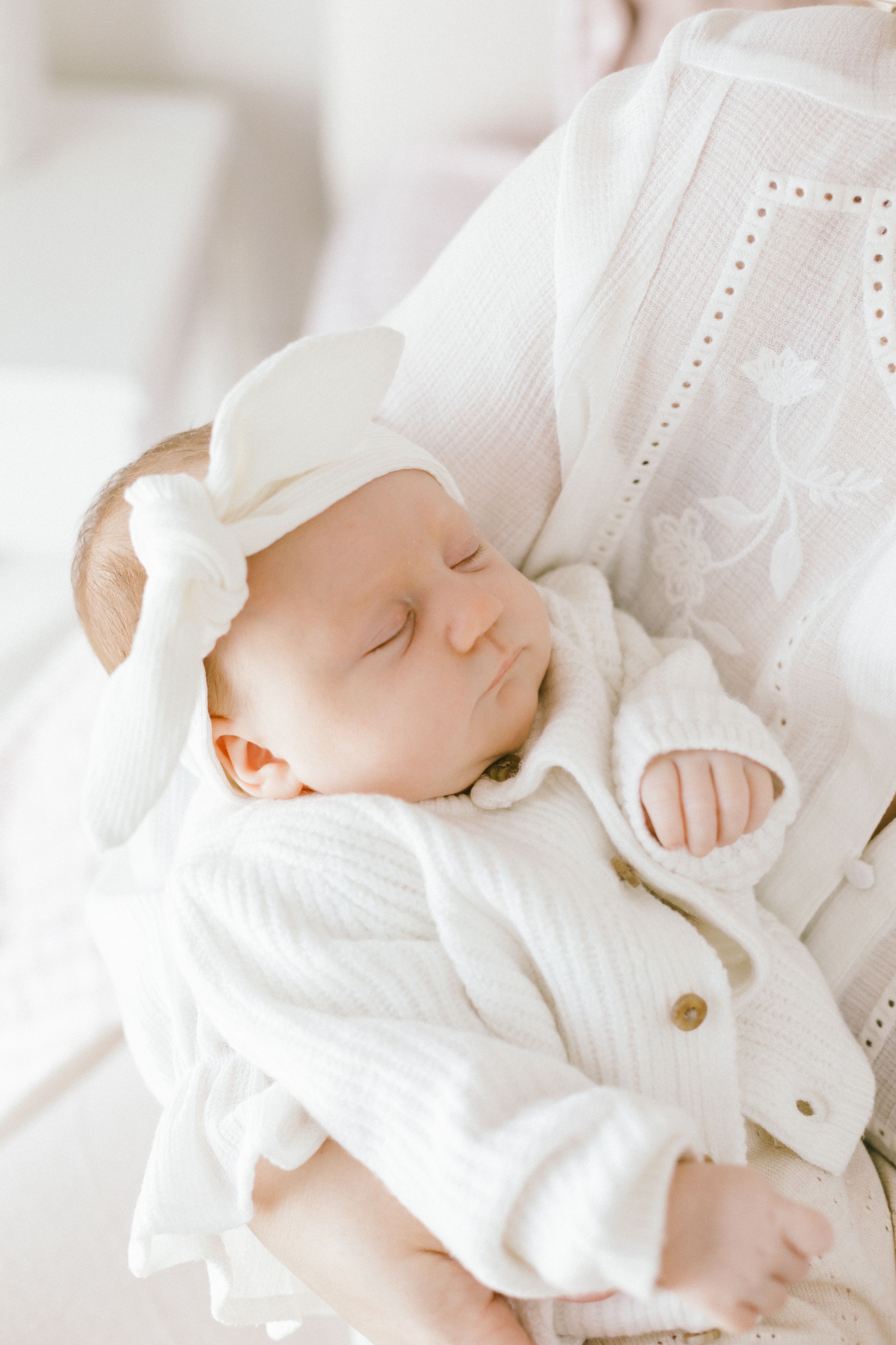 Sleeping newborn baby in white outfit and bow headband cradled in mother's arms during newborn photography session.