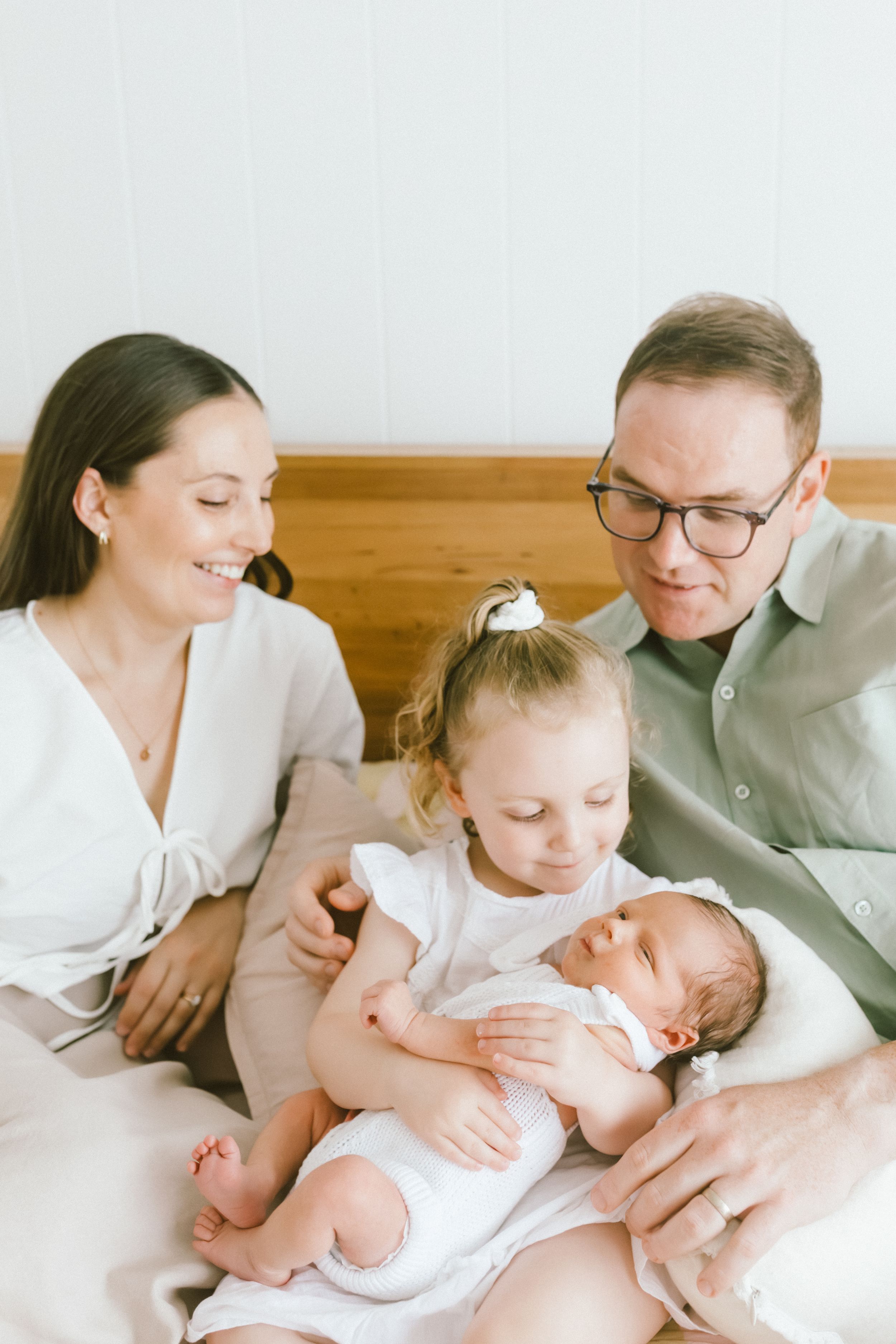 Happy family of four sits on a bed, parents smiling as toddler girl gently holds their newborn baby sibling.