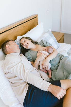 New parents lying on bed together, mother holding newborn baby while father looks on lovingly.