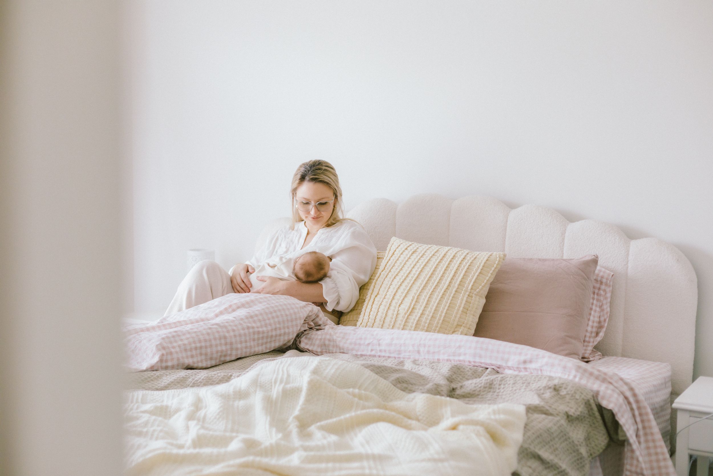 Mother breastfeeding newborn baby while sitting on a cozy white bed with soft pink pillows and blankets.