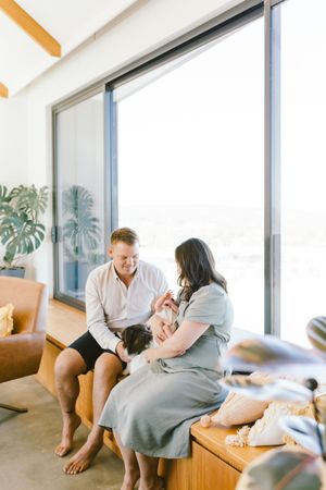 A couple sits by large windows holding their newborn baby, with a dog nearby, in a bright modern home.