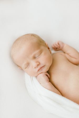 Peaceful newborn baby sleeping wrapped in white swaddle blanket on soft white background.