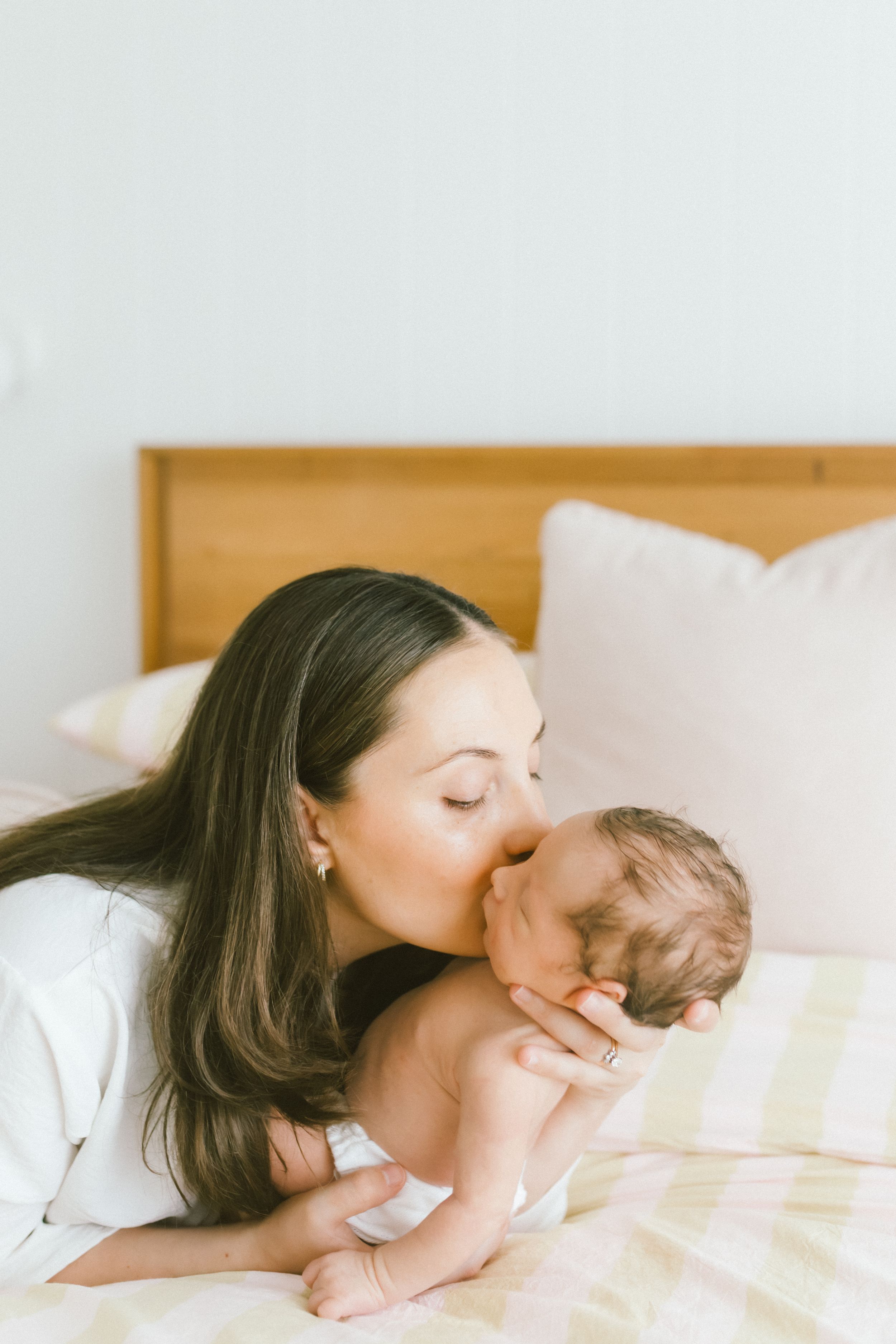 A mother with long dark hair gently kisses her newborn baby while lying on a bed with white pillows.