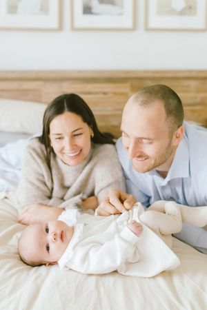 Happy parents lying on bed smiling at their newborn baby dressed in white.