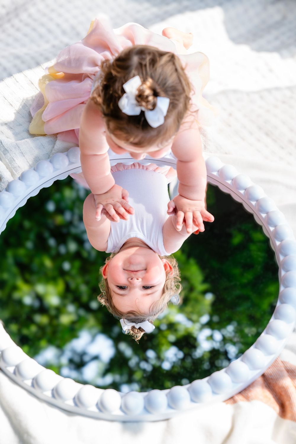 Baby wearing pink dress and bow reflected in ornate white circular mirror against green background.