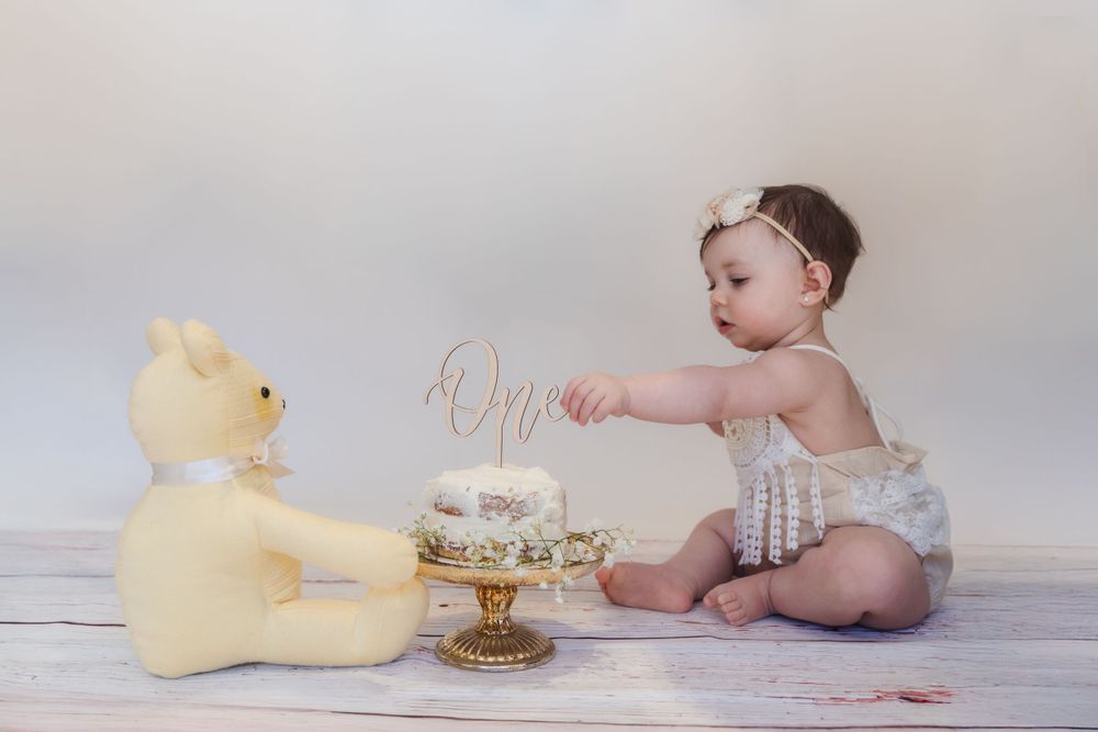 A baby in white sits next to a yellow teddy bear with a small cake for a first birthday photoshoot.
