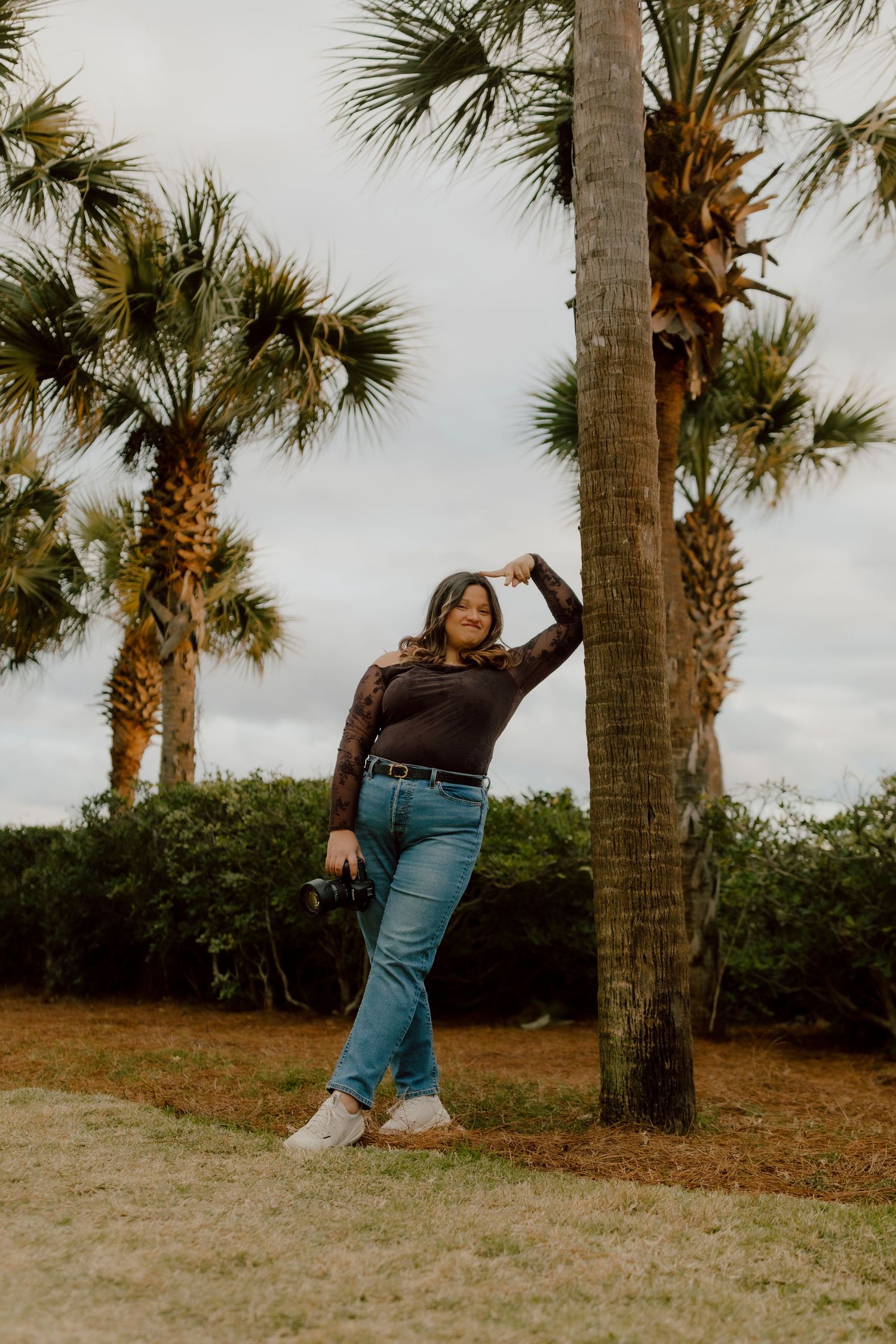 Smiling woman in dark lace top and jeans posing by palm tree in coastal park setting.