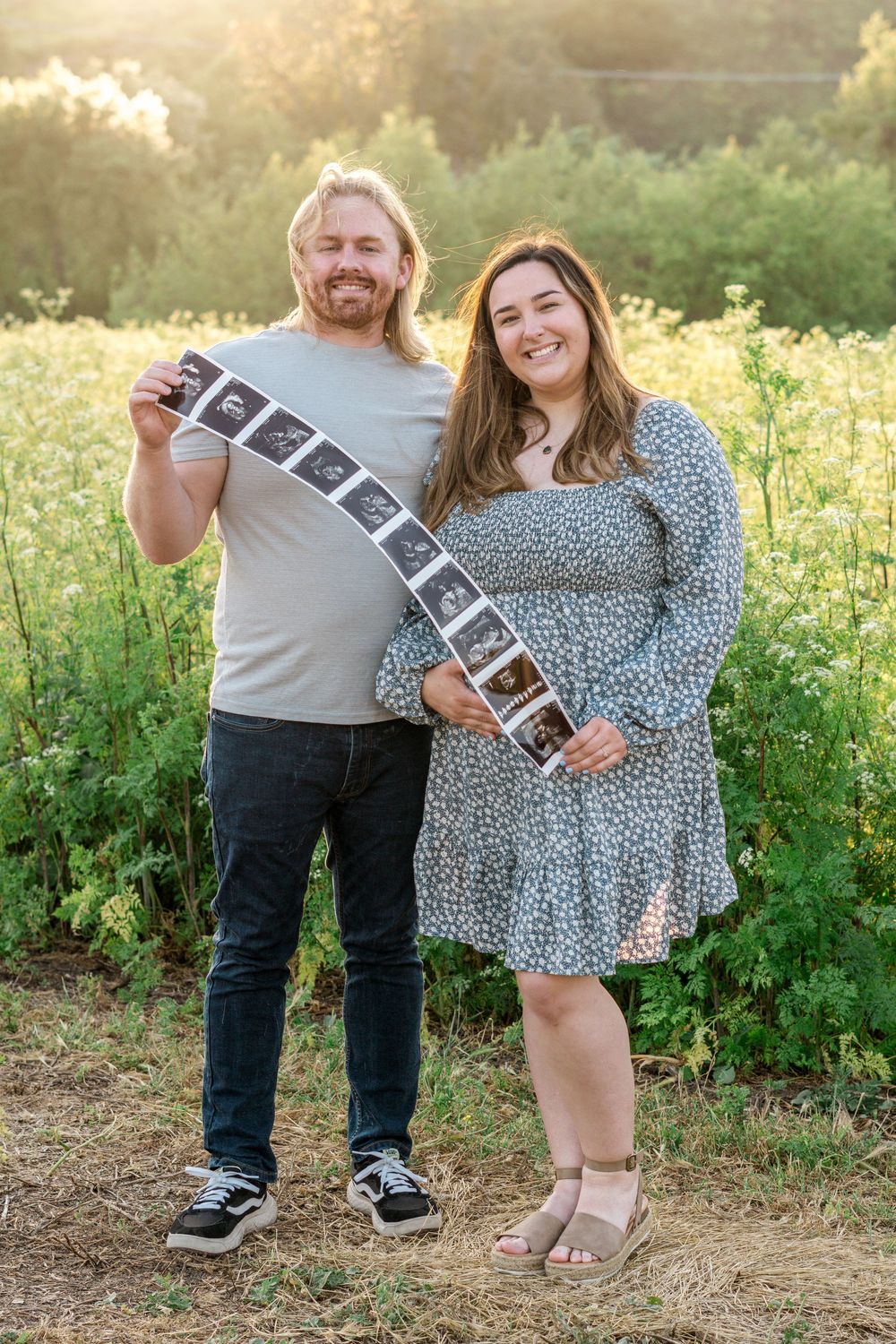 Couple holding ultrasound photos in a golden sunlit field during pregnancy announcement photoshoot.