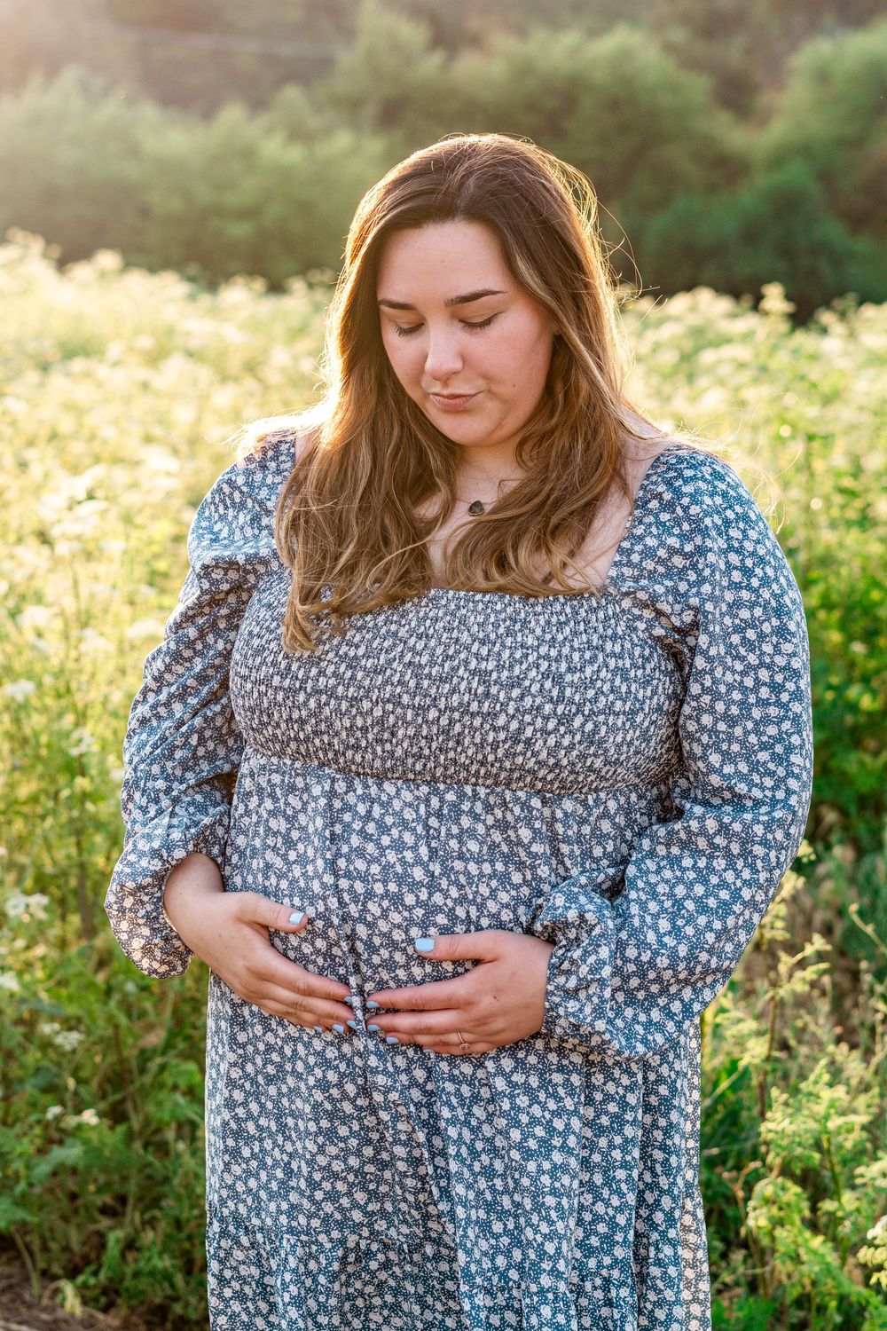 A person in a floral print dress stands in a sunlit meadow at golden hour, hands resting gently on their stomach.