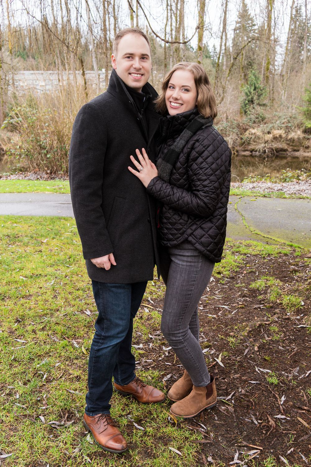 A couple in winter coats embraces while standing on a path surrounded by bare trees and grass.