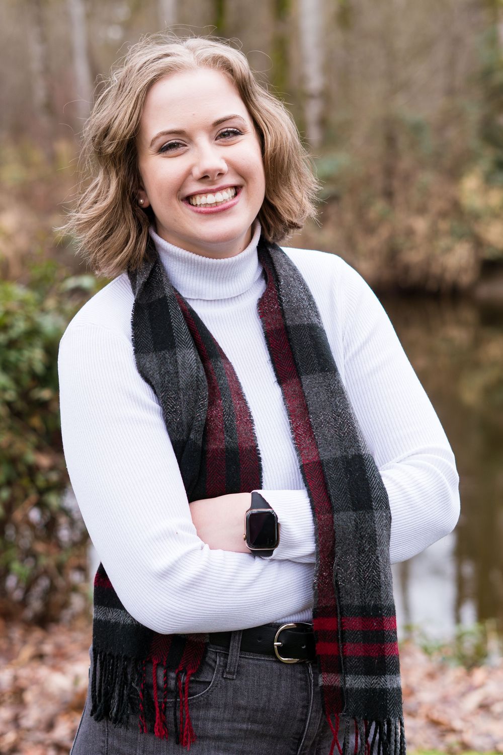A person wearing a white turtleneck sweater and plaid scarf poses outdoors in autumn.
