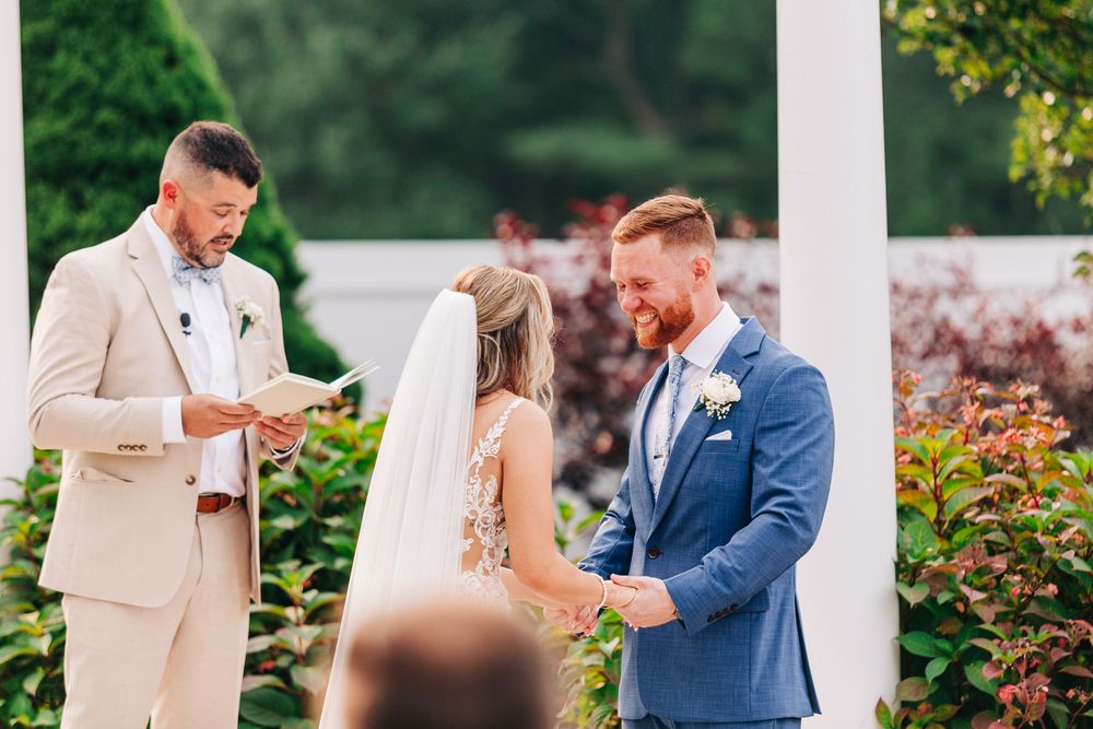 A couple exchanges vows during an outdoor wedding ceremony with a white pillar and garden backdrop.