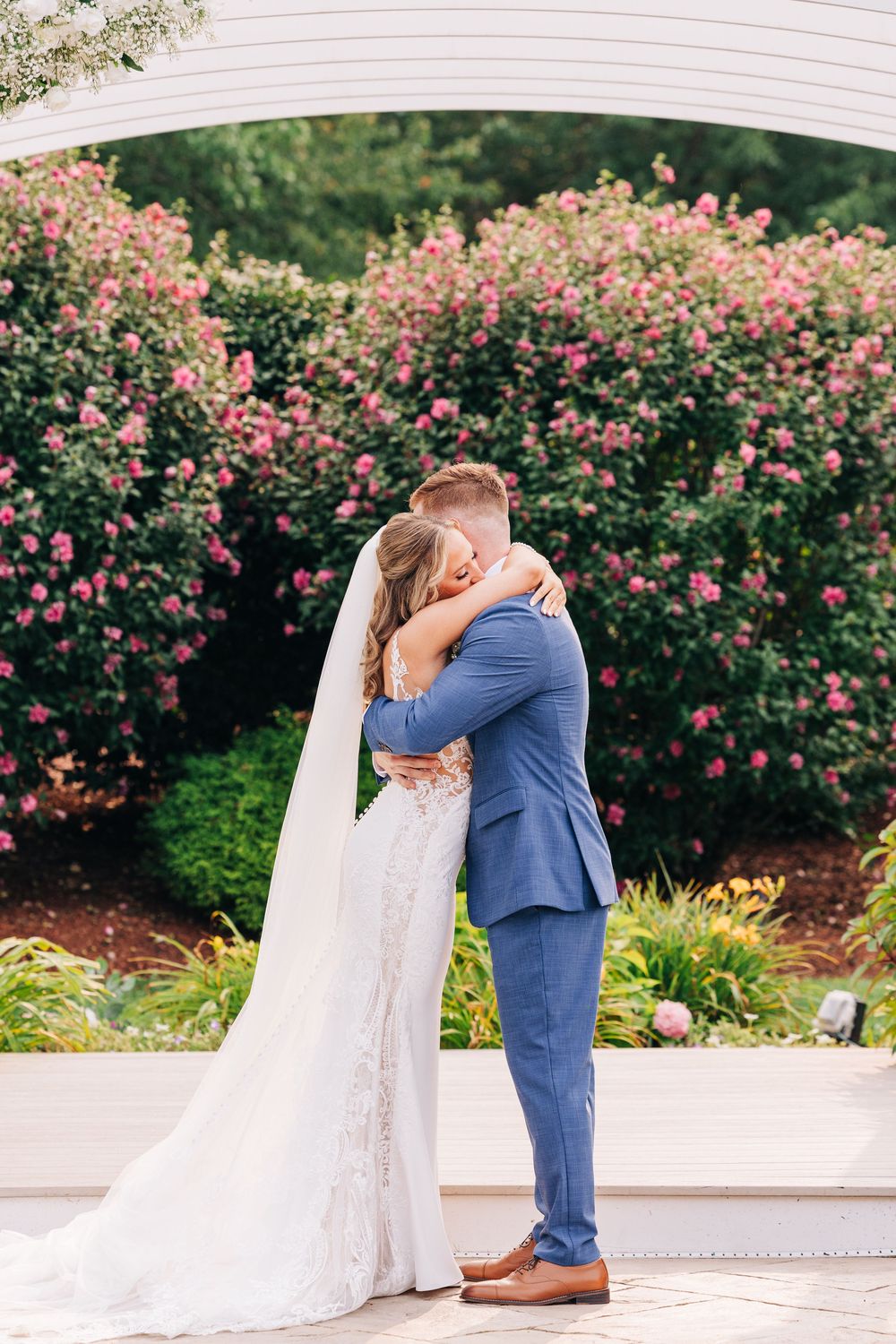 Newlywed couple shares romantic kiss in garden setting with blooming pink rose bushes and white pergola.
