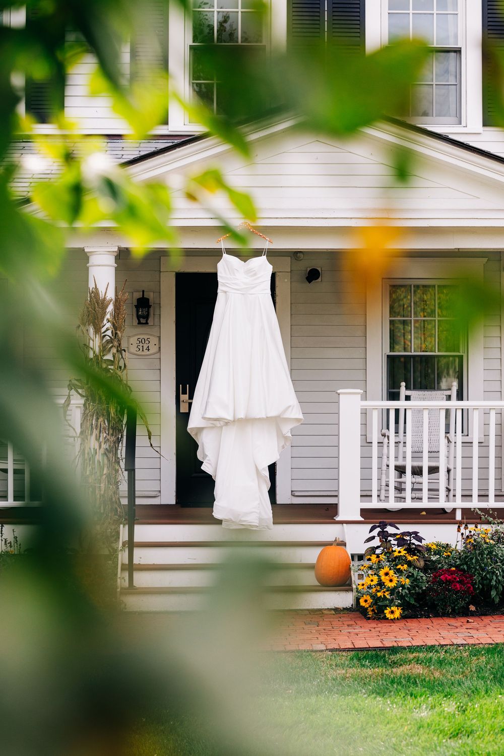 White wedding dress hanging outdoors against classic white farmhouse porch with autumn pumpkin and floral decor.