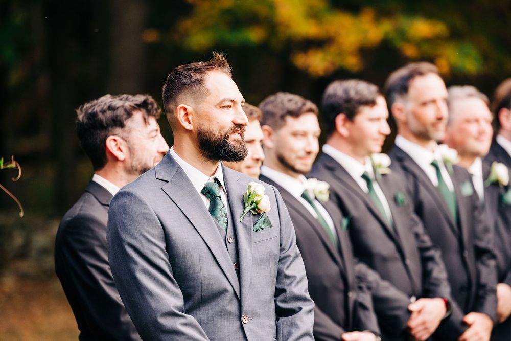 Groomsmen in dark suits stand in line at an outdoor wedding ceremony with autumn foliage in the background.