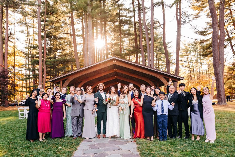 Large wedding party poses together outdoors in a pine forest at sunset with warm golden light filtering through trees.