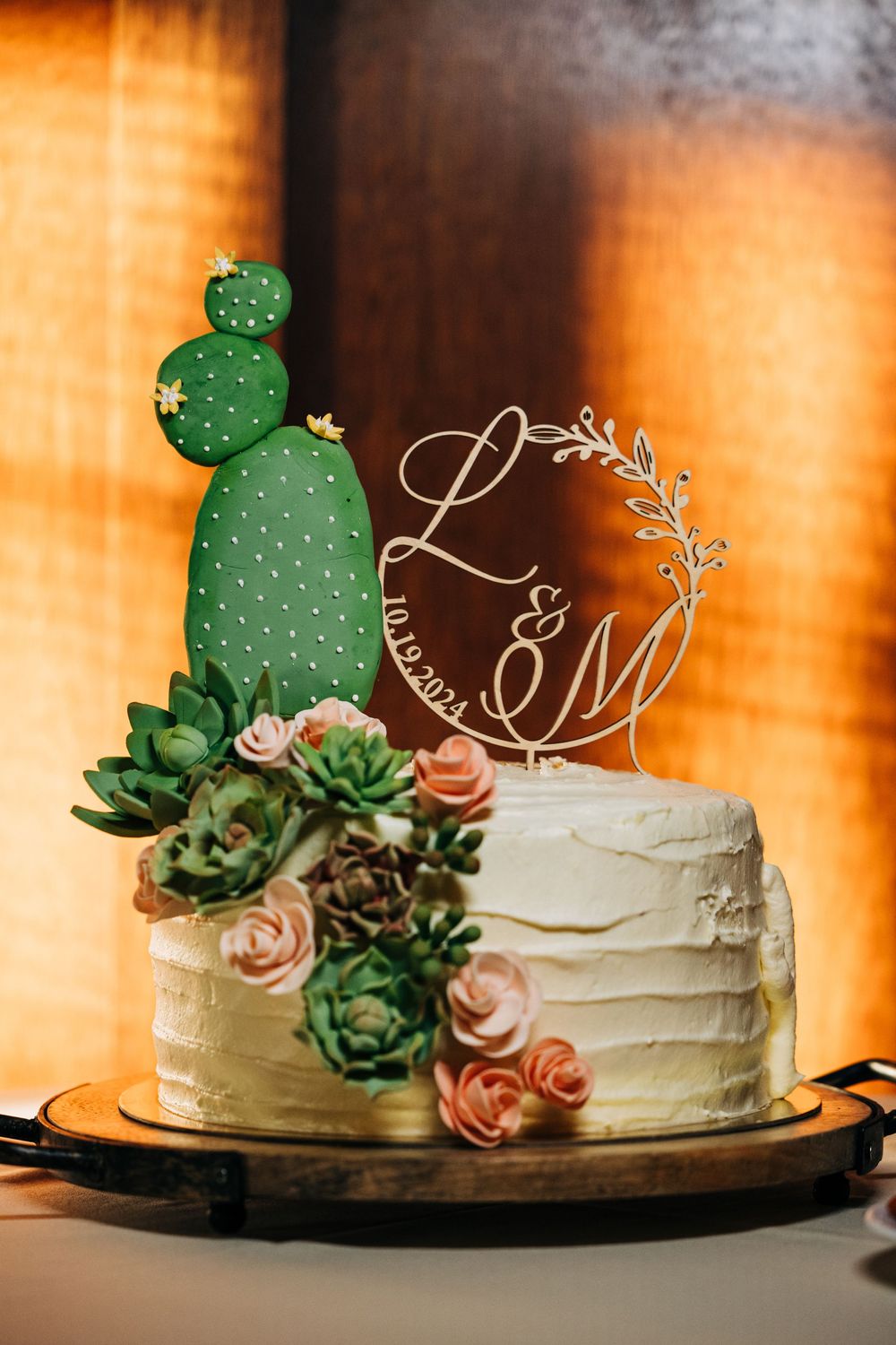 Wedding cake decorated with green succulents and peach roses on a dark background with warm lighting.