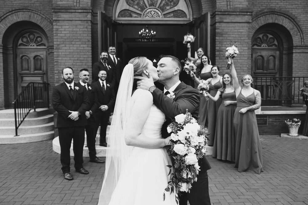 A wedding party poses on brick steps in front of an ornate historic building with arched doorways.