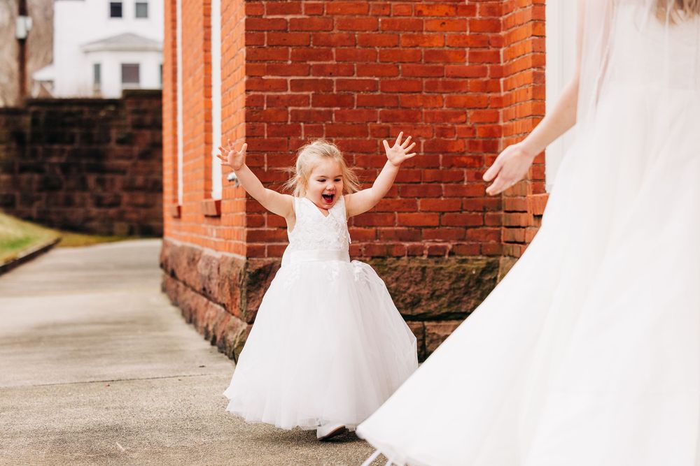 A person in a white dress twirls joyfully in front of a red brick wall.