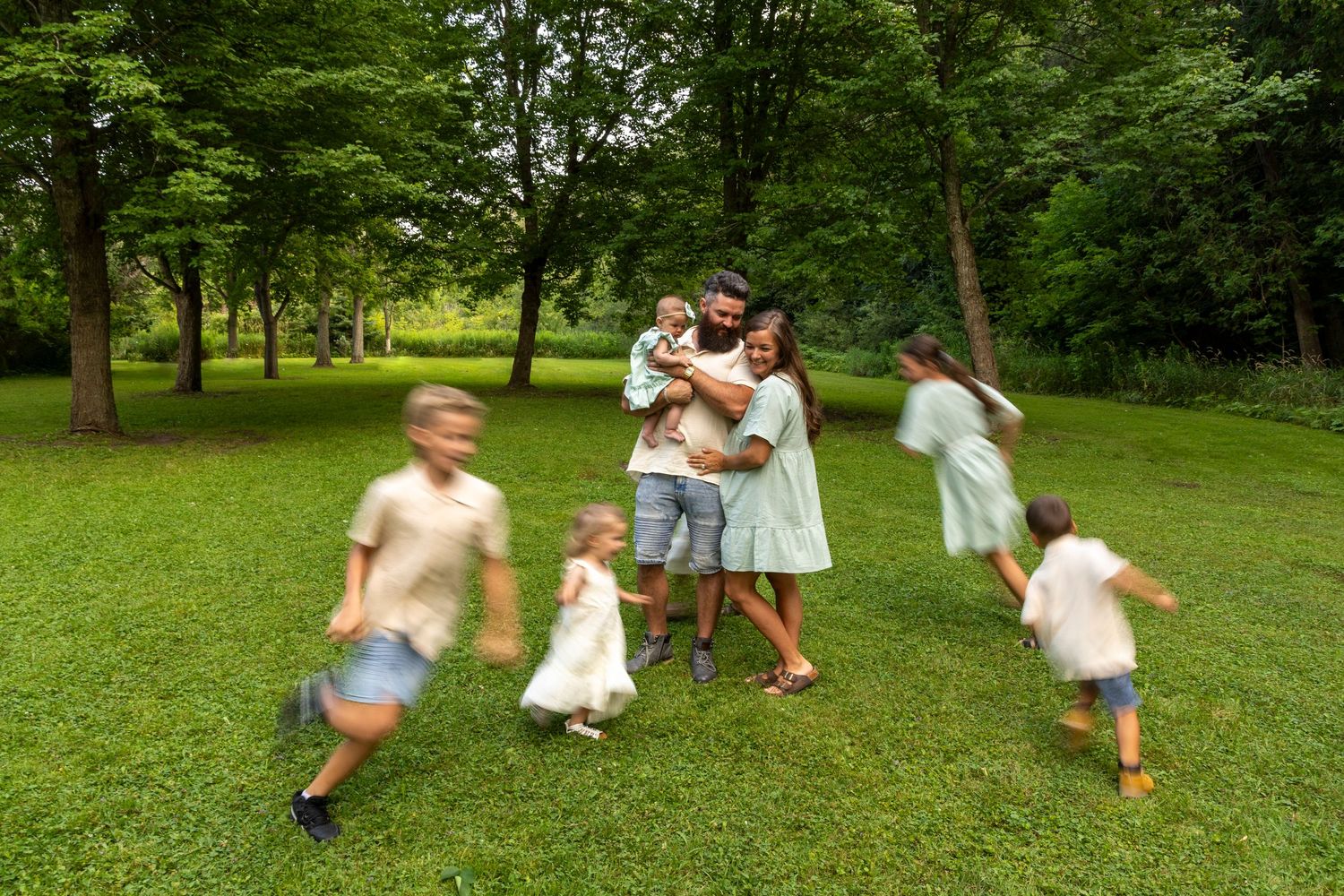 A mom, dad and baby hugging in a park with their 5 additional children running around them (blurred)