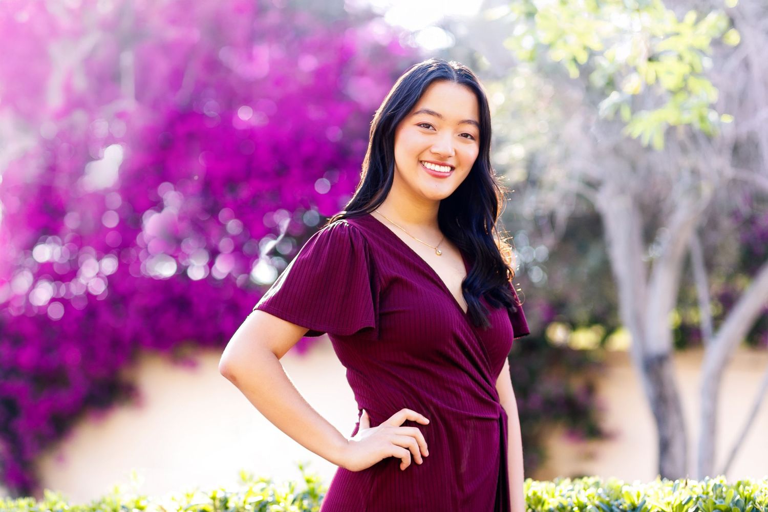 A high school senior in a white summer dress poses outdoors at golden hour with sunlit trees creating long shadows.