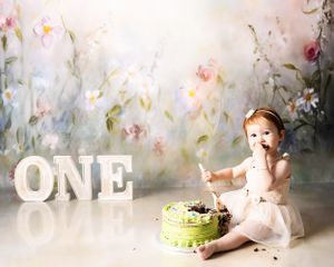 A green birthday cake sits beside glowing 'ONE' letters against a dreamy floral backdrop for a first birthday celebration.