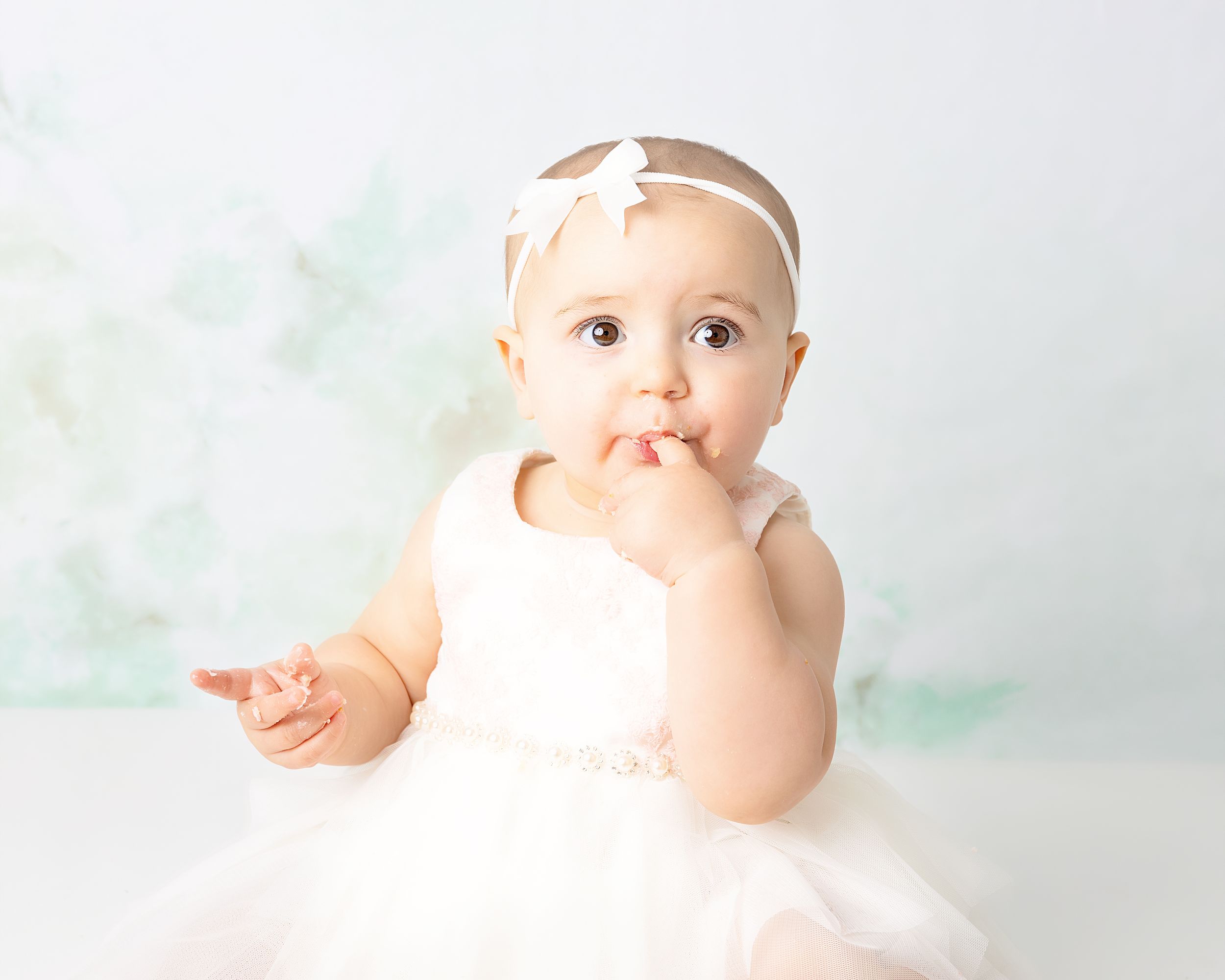 Baby in white tulle dress and headband sits against pastel background while sucking thumb.