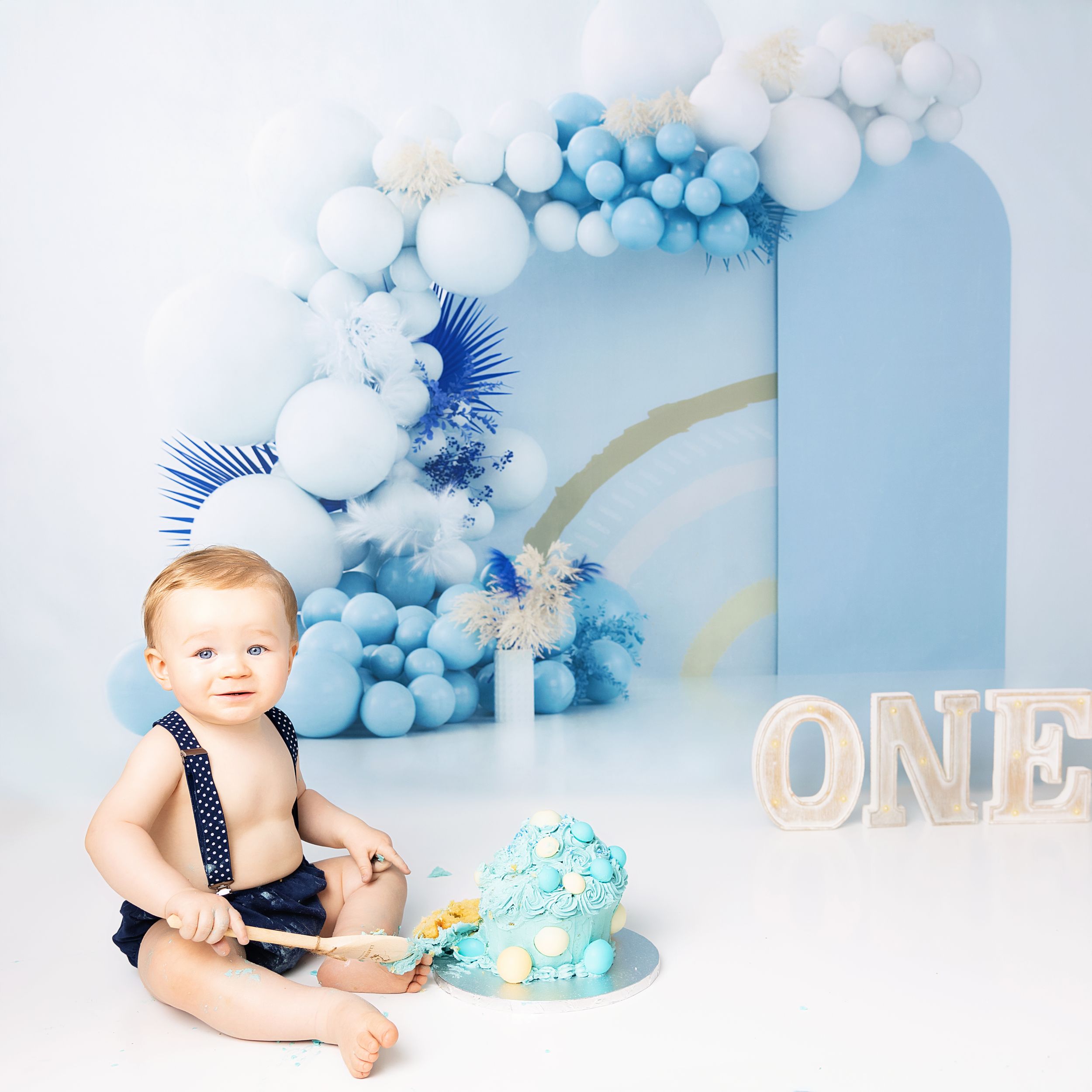 A festive blue and white balloon arch backdrop with light decorations and 'ONE' letters for a first birthday celebration.