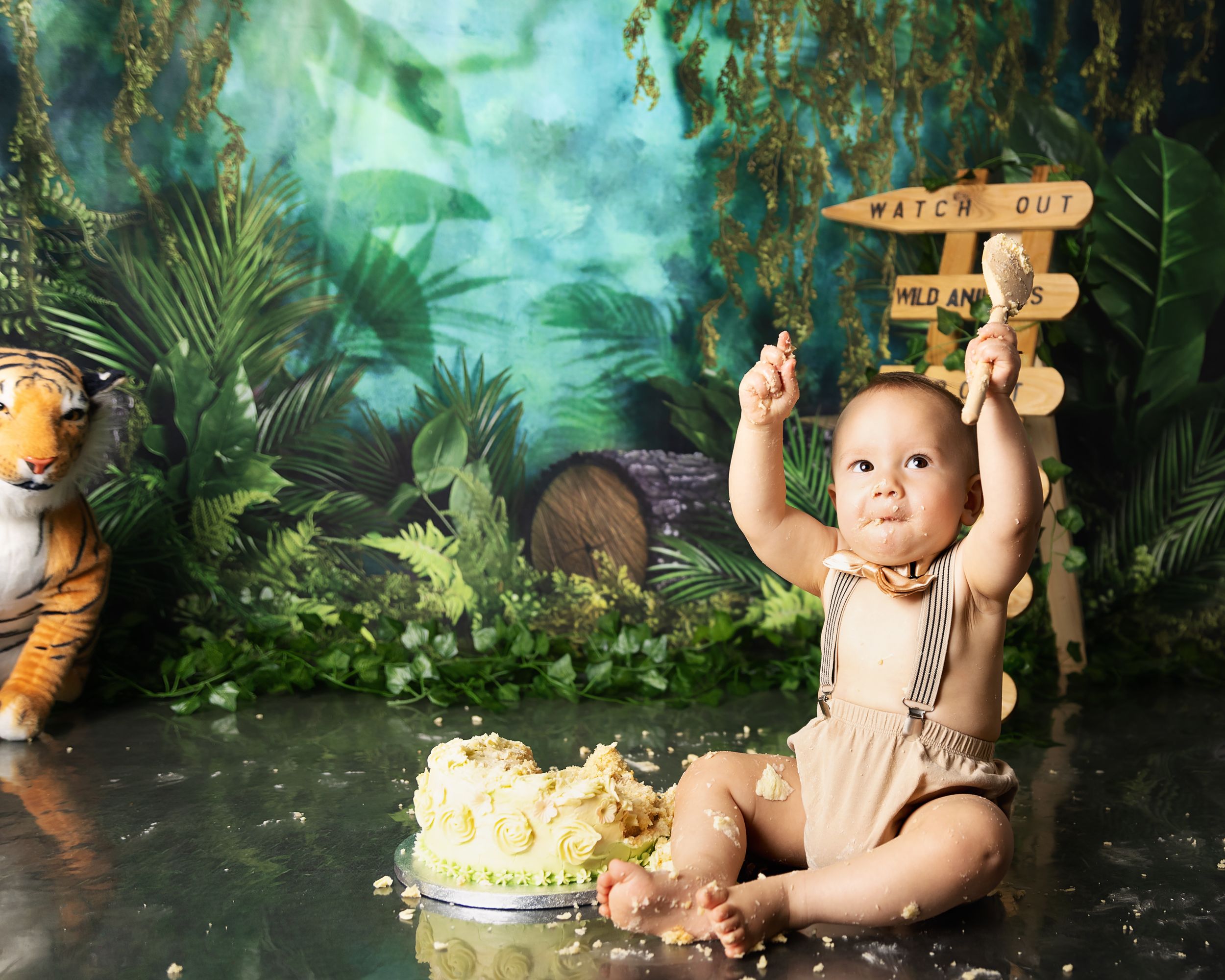 A baby in a romper celebrates their first birthday with cake in a jungle-themed photography setup with props and decorations.