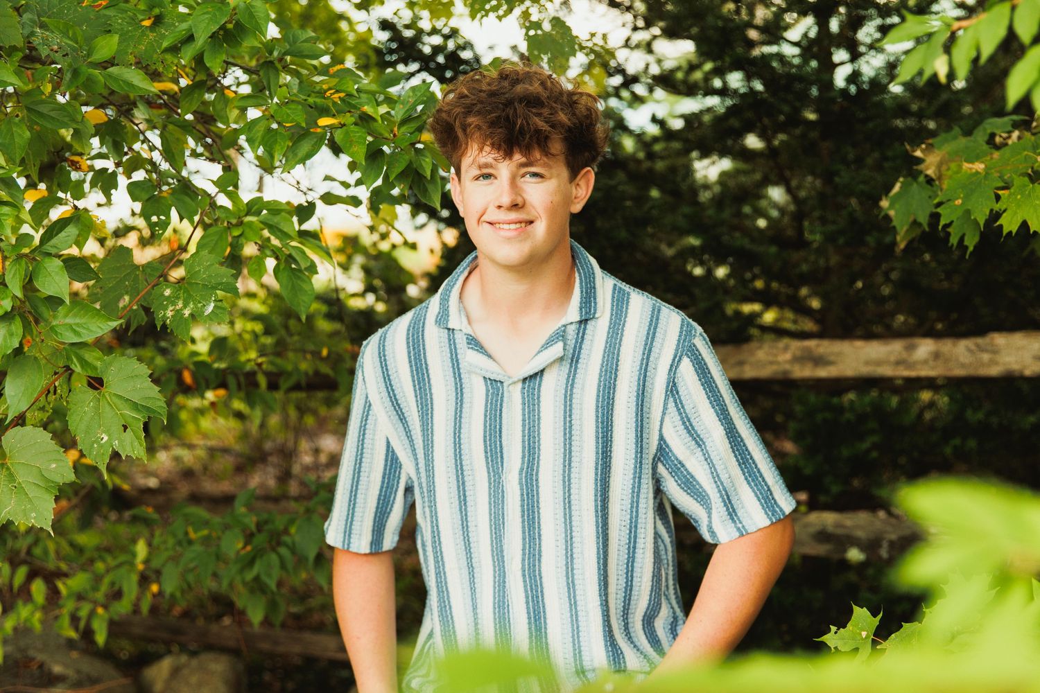 Smiling teenage boy in striped blue shirt standing outdoors among green trees and wooden fence.
