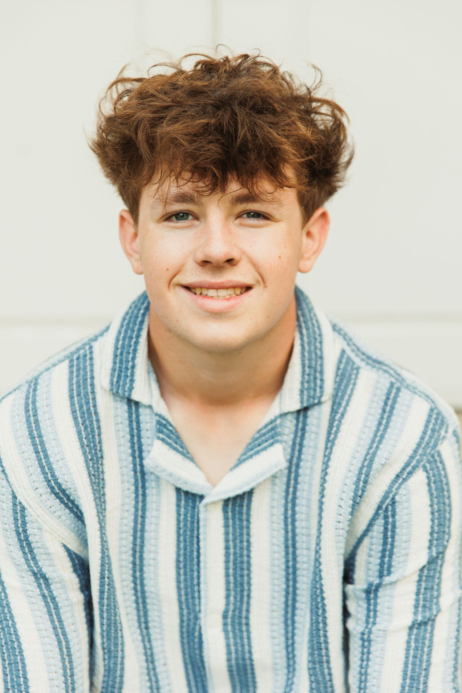 Smiling teenage boy with curly brown hair wearing a blue and white striped shirt against a light gray background.