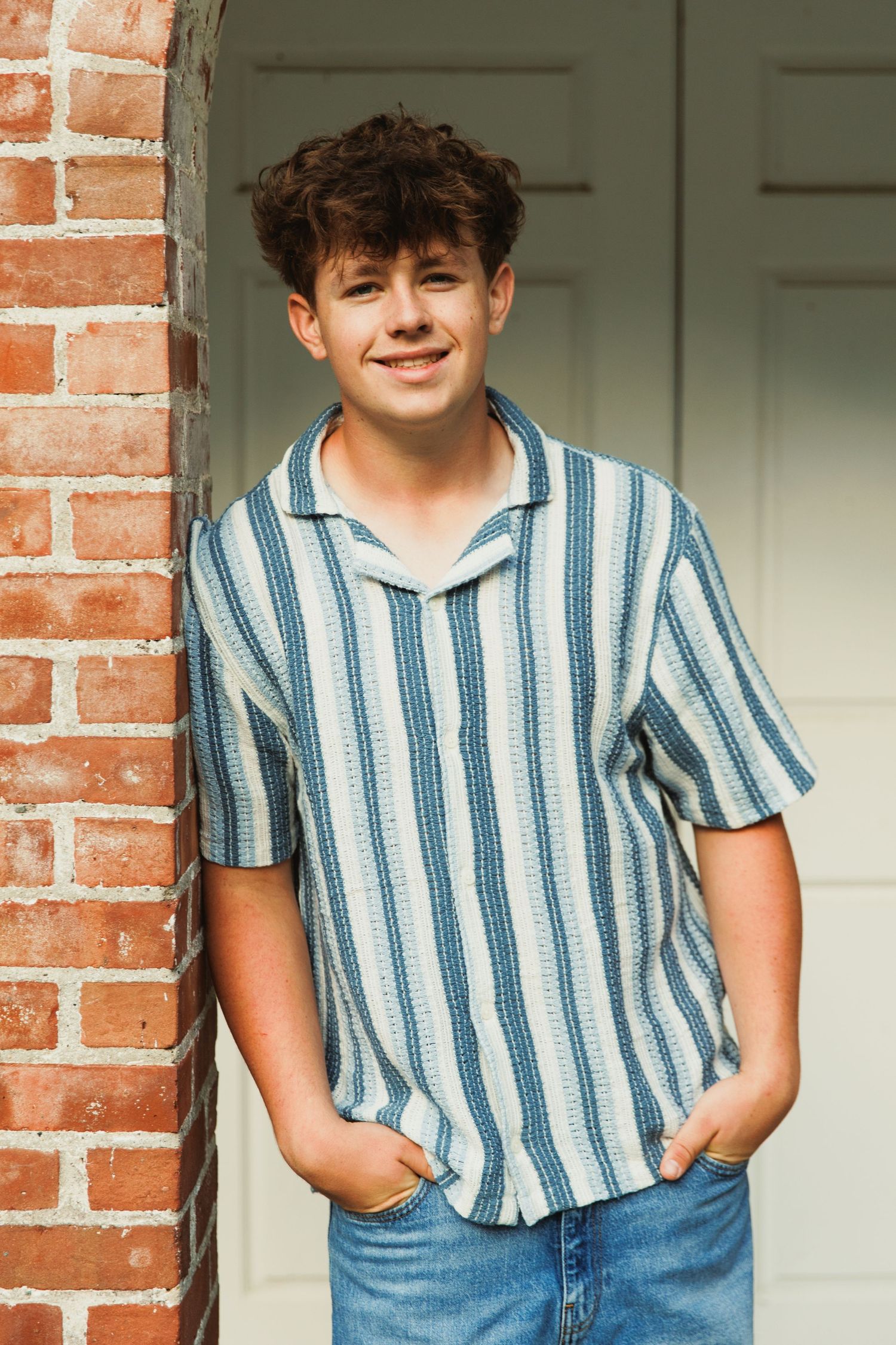 A smiling teenage boy in a striped blue shirt and jeans leans against a brick column outdoors.