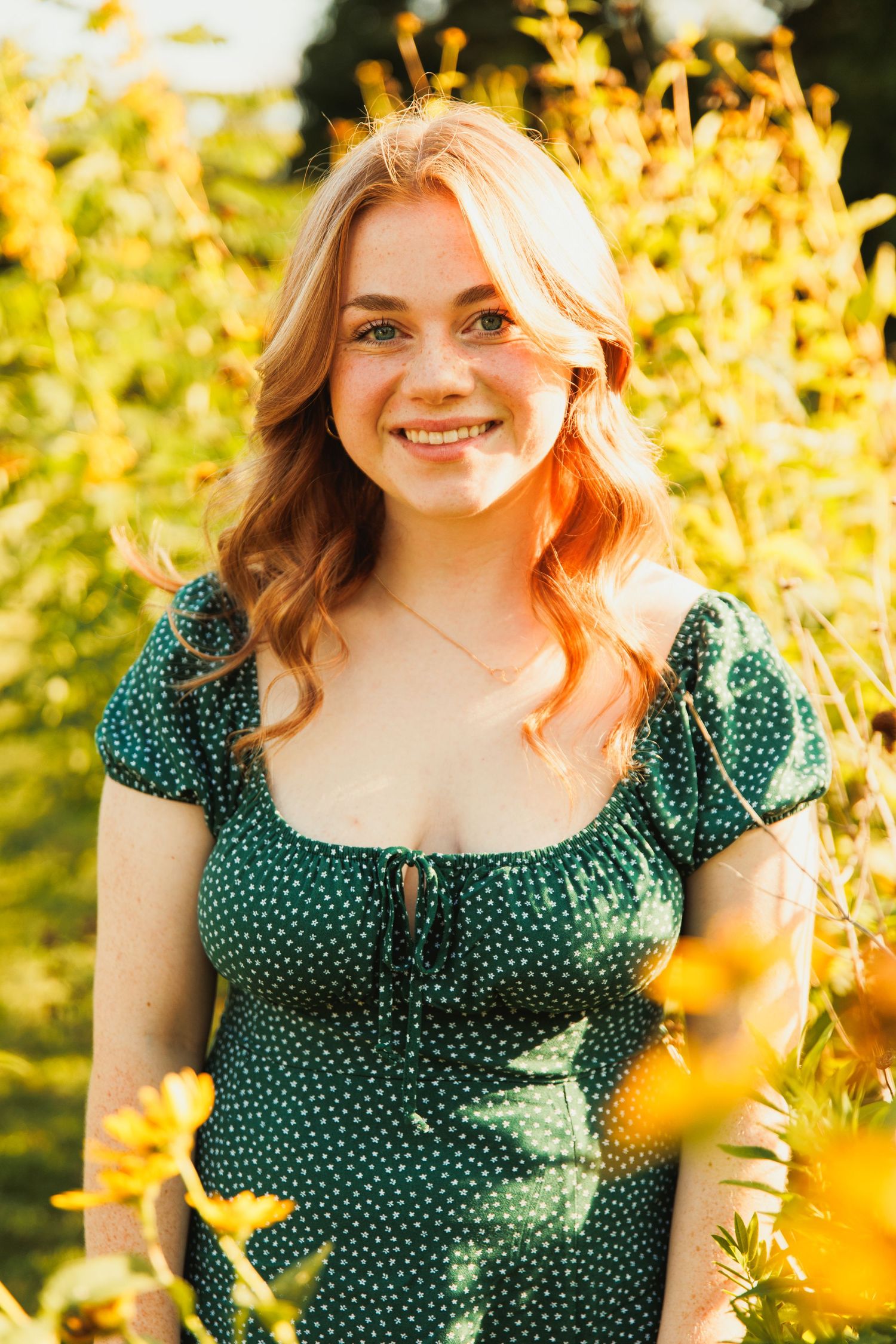 Smiling young woman with red hair in green polka dot dress standing among yellow wildflowers outdoors.