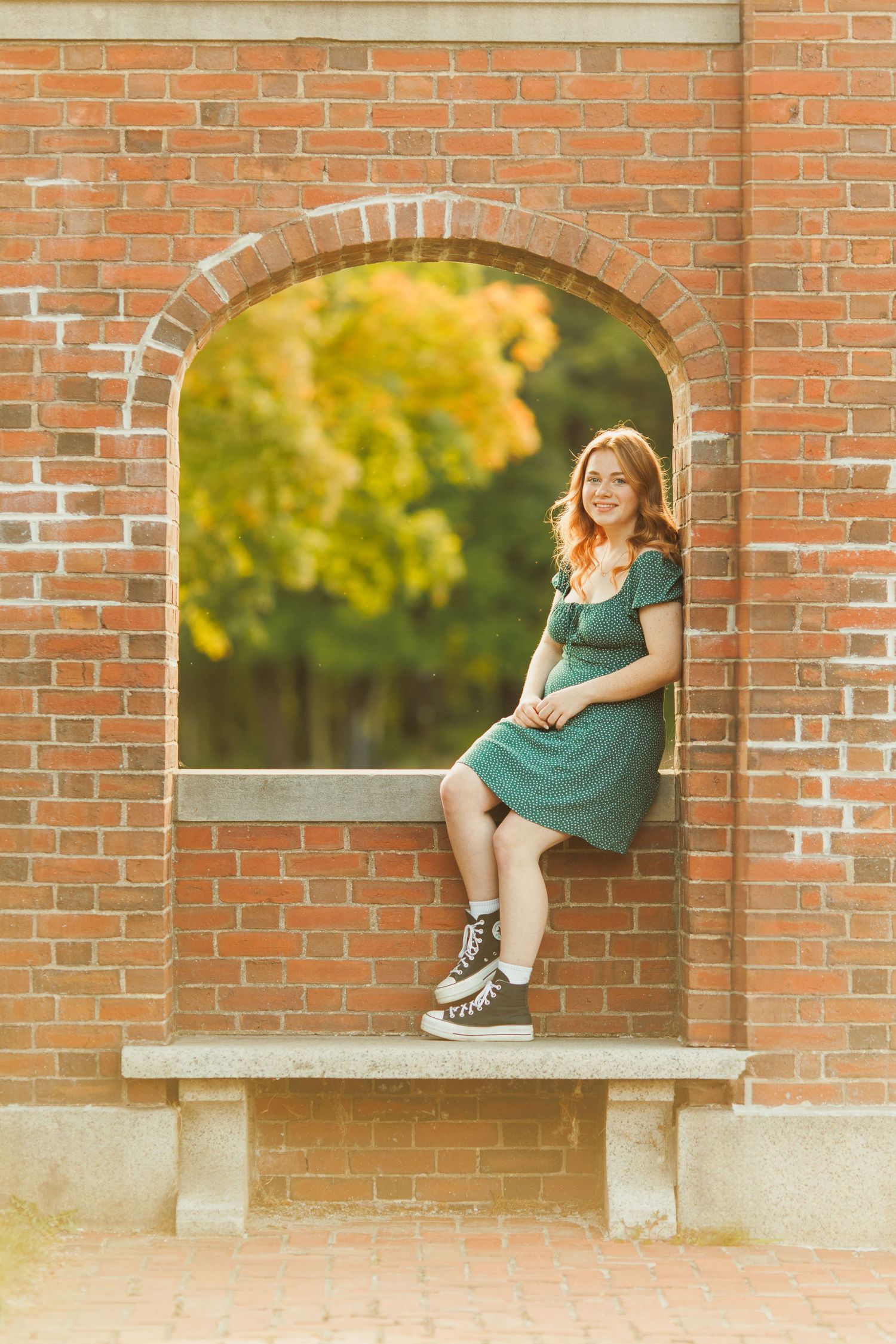 Young woman in green dress and sneakers sitting in a brick arch window with autumn trees in background.