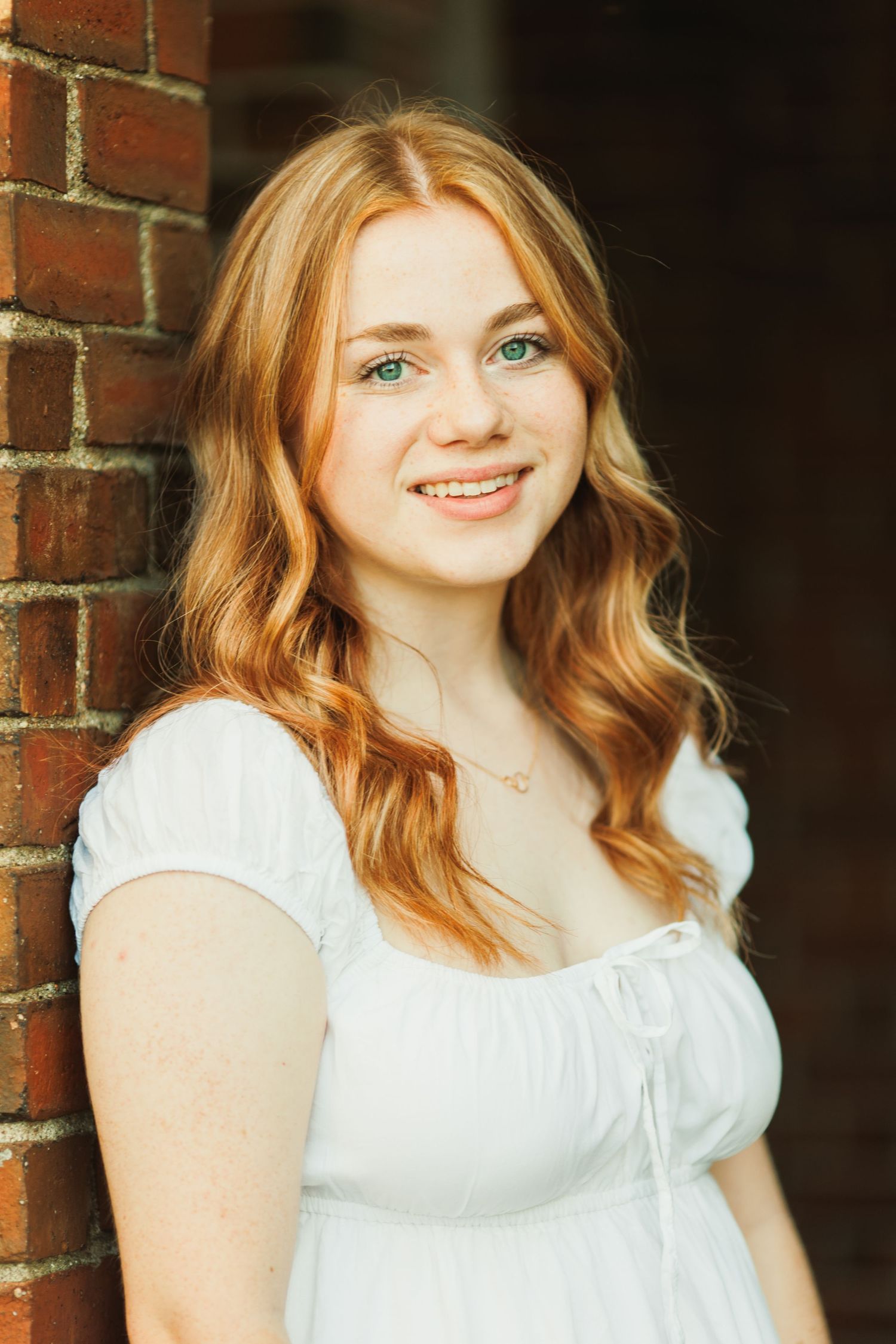 Young woman with red wavy hair and green eyes smiling in white dress leaning against brick wall.