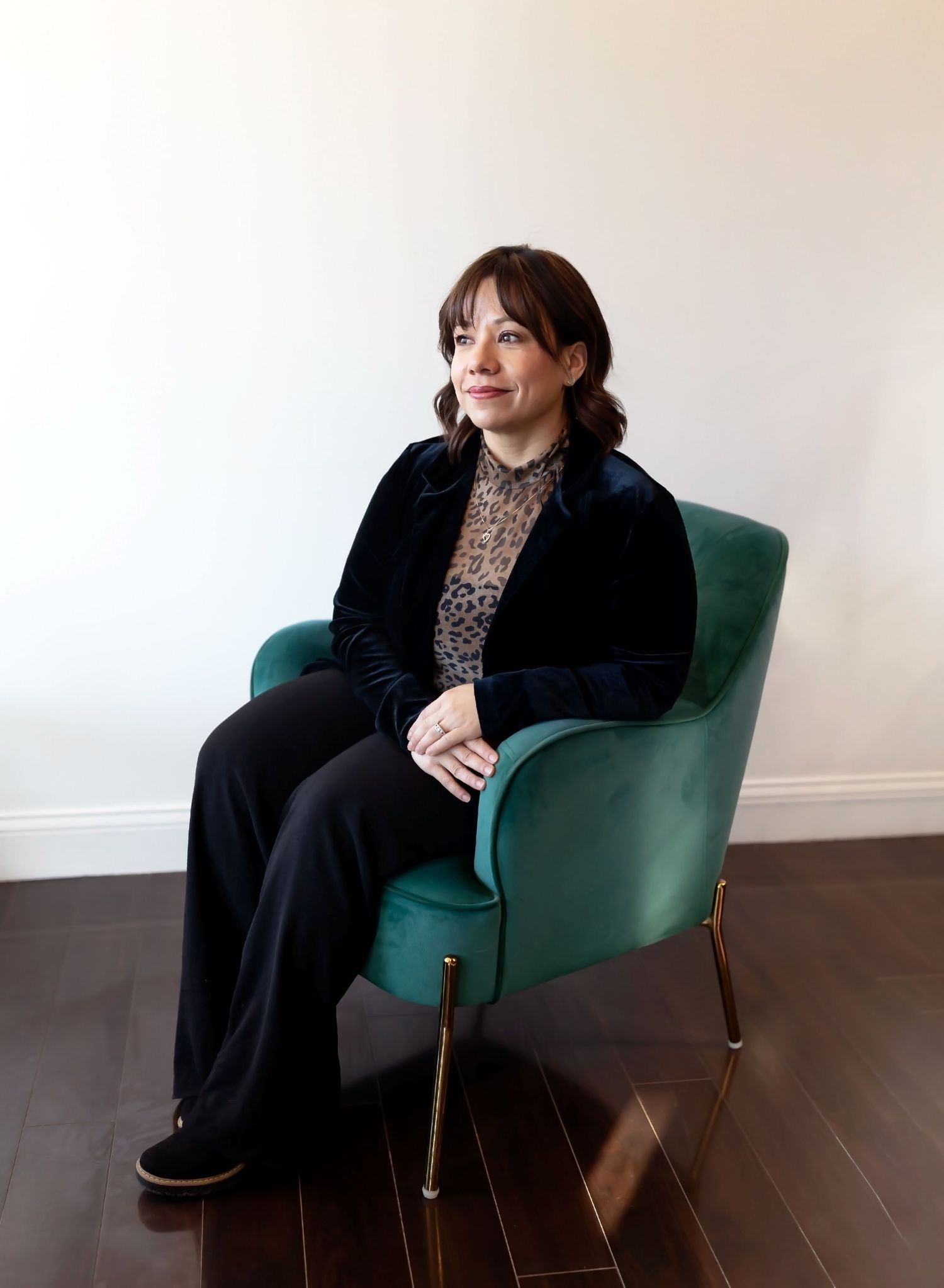 Person in black attire sits thoughtfully in a modern emerald green armchair against white wall. Branding photo session of a hair stylist