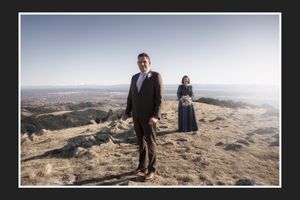 A couple poses on a rocky hilltop, man in suit and woman in blue dress, with vast landscape views behind them.