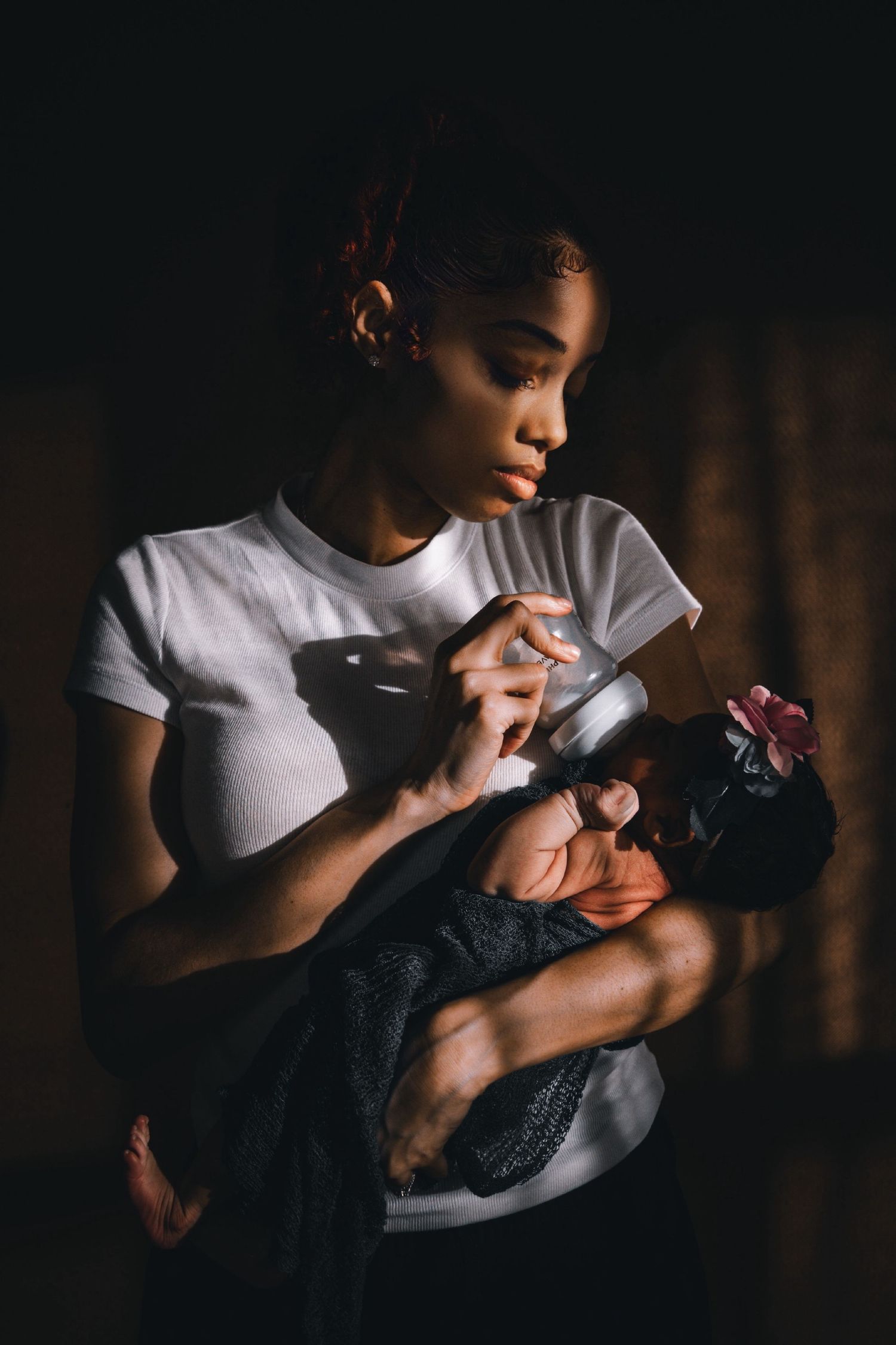 Person in white t-shirt holds and feeds an infant in a dark, intimate setting.