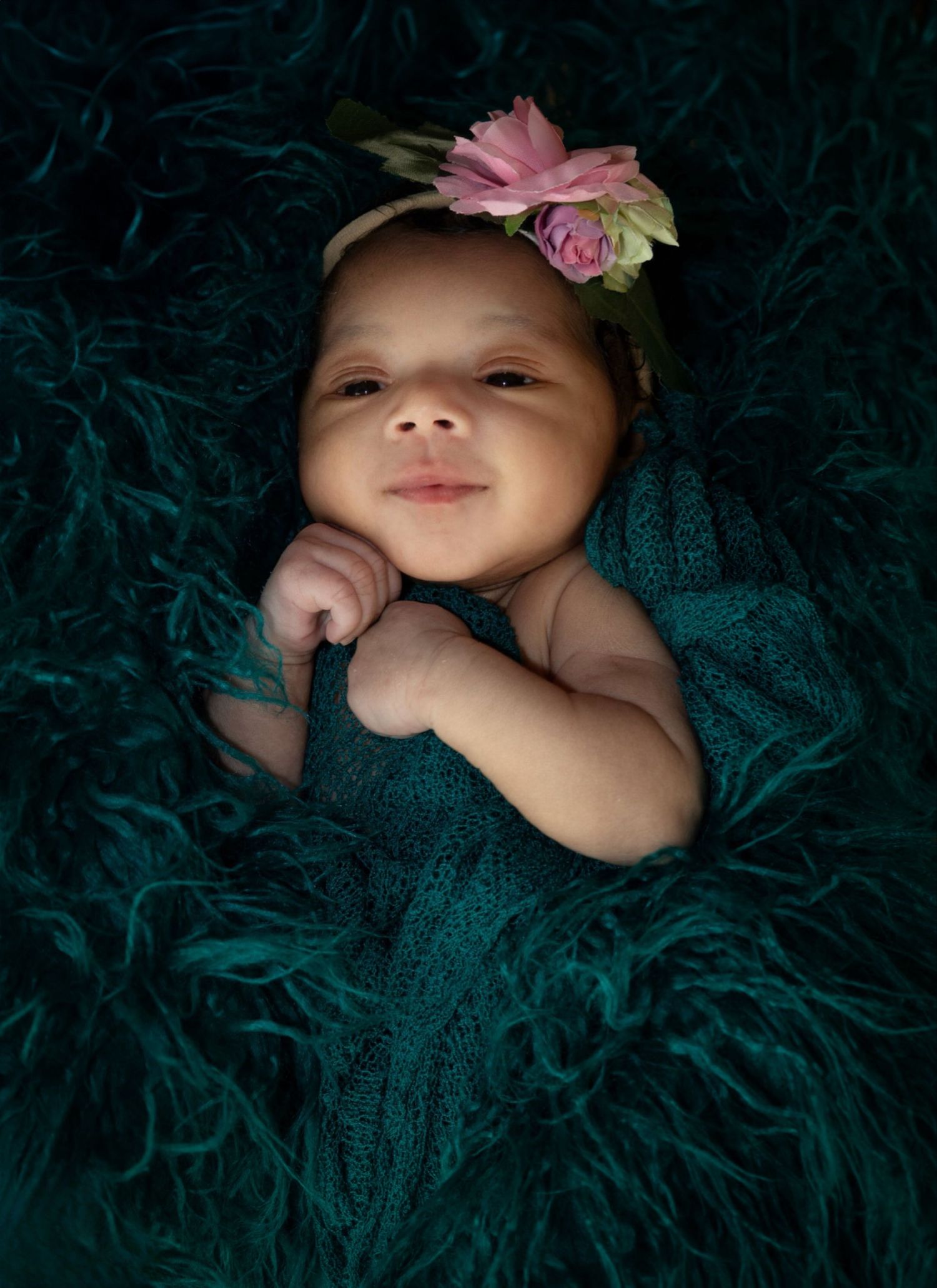 Baby wearing floral headband lies on teal textured fabric in professional portrait.