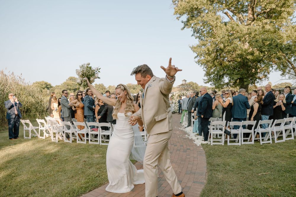 Outdoor wedding ceremony setup with white chairs and guests gathering on a sunny hillside.