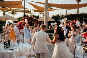 Wedding guests dance and celebrate at an outdoor reception under white canopy tents during sunset.