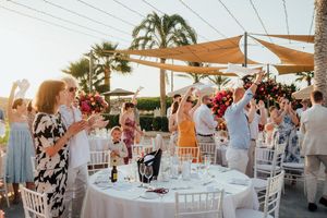 Wedding guests celebrate during an outdoor reception under shade sails at sunset with palm trees in the background.