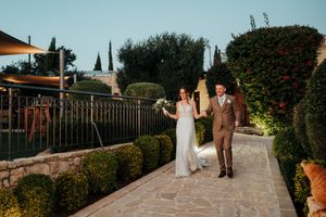 A couple walks along a scenic stone path surrounded by lush gardens at a Mediterranean-style venue during sunset.