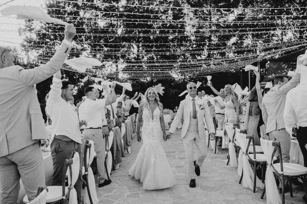 Wedding guests form an archway with raised arms as a couple in white walks beneath at an outdoor celebration.
