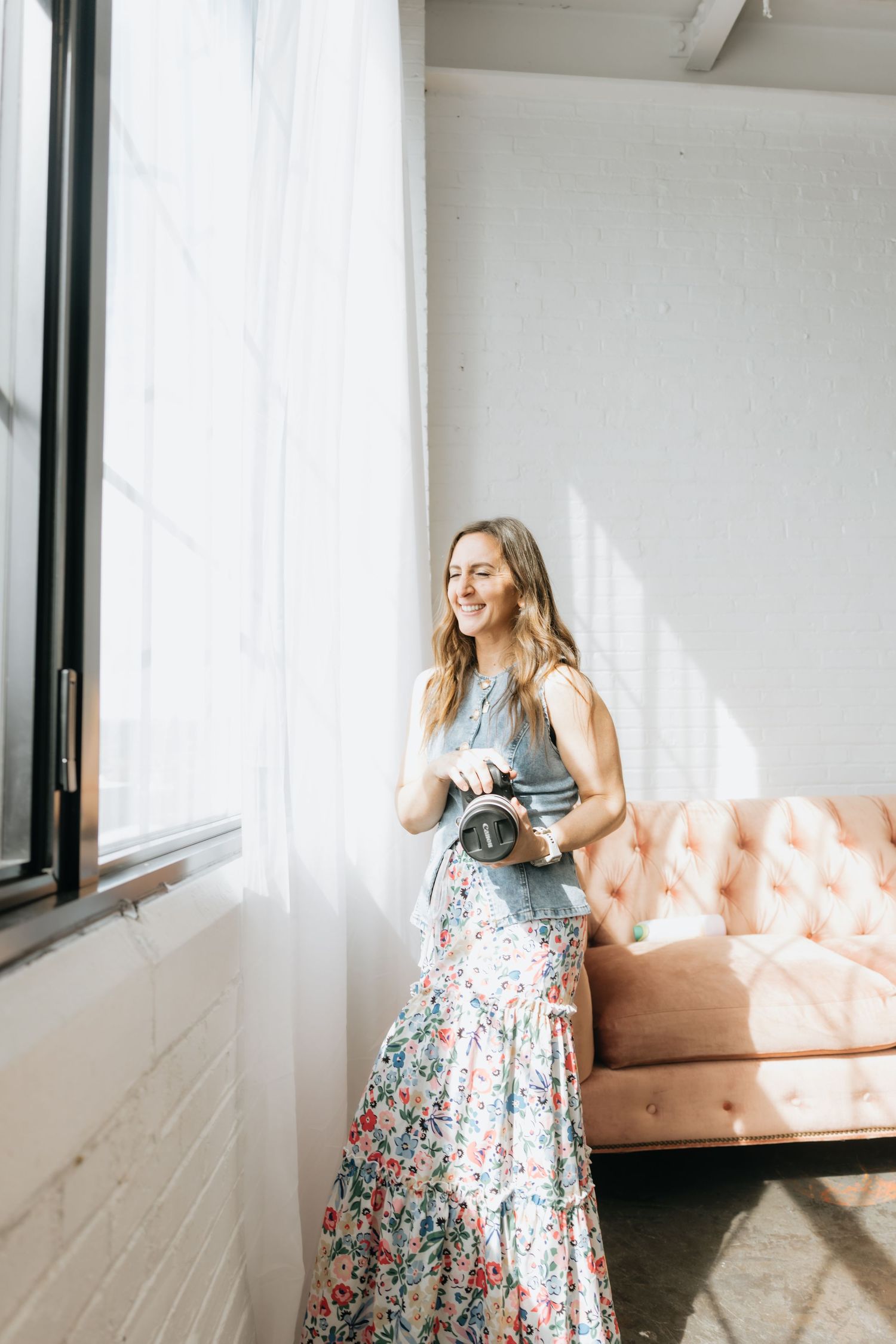 A smiling woman holds a camera while standing by a large window in a bright, airy studio space.